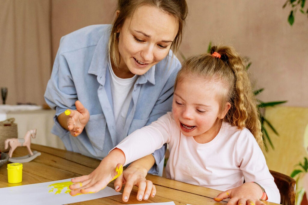 An adult assists a child with Down Syndrome finger paint