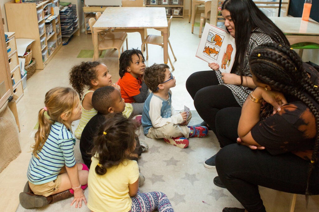 Two teachers read a book to young children circled on the floor before them