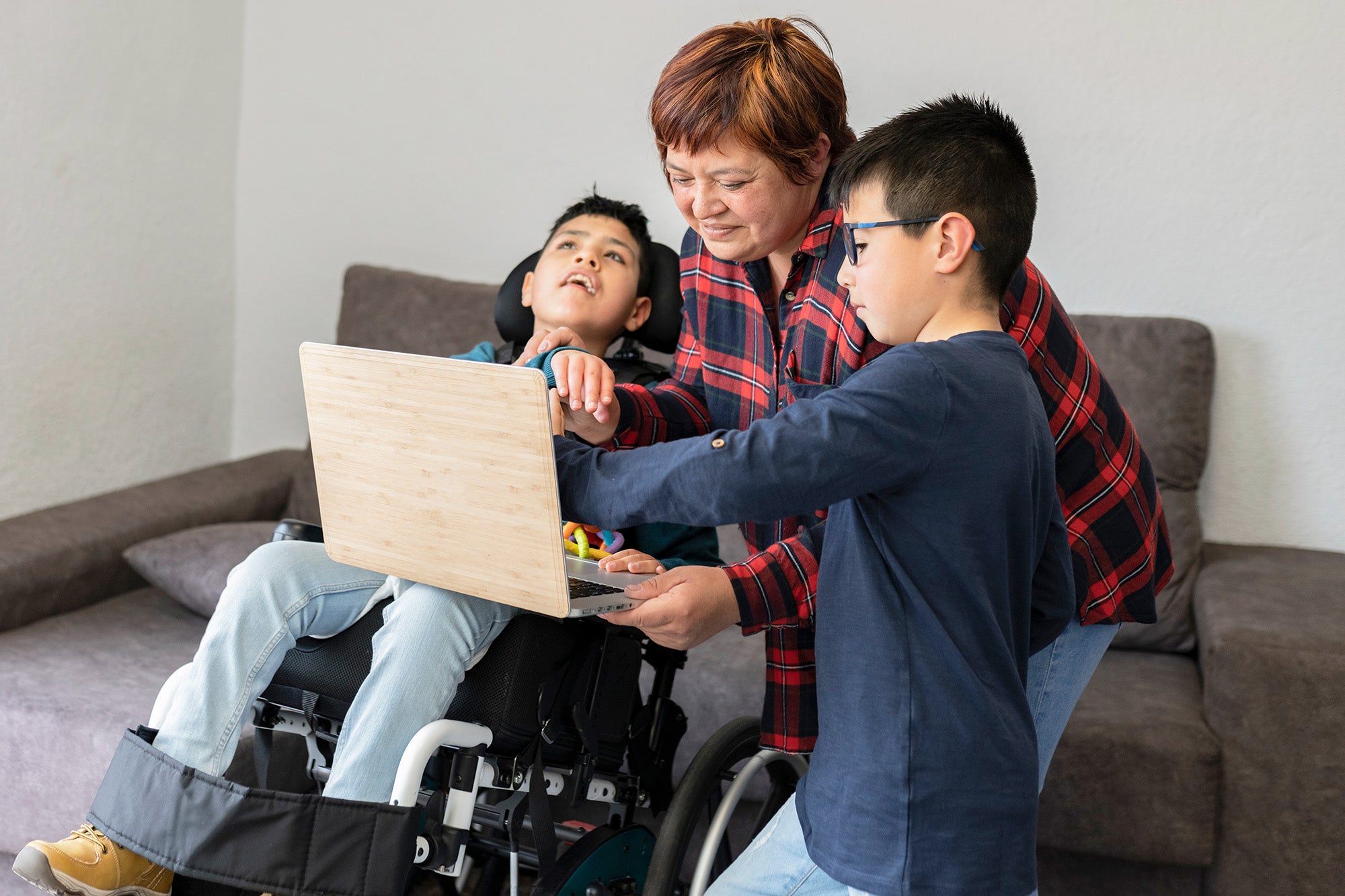 A young family member assists a boy in a wheelchair with a laptop as an adult caregiver looks on