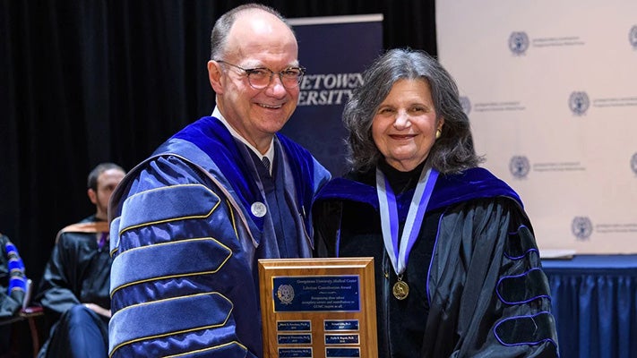 Phyllis Magrab and Jack DeGioia wearing academic regalia stand together holding a plaque