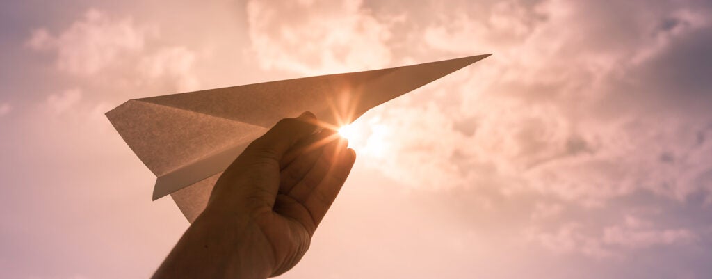 A hand holds a paper plane up in a sunlit sky