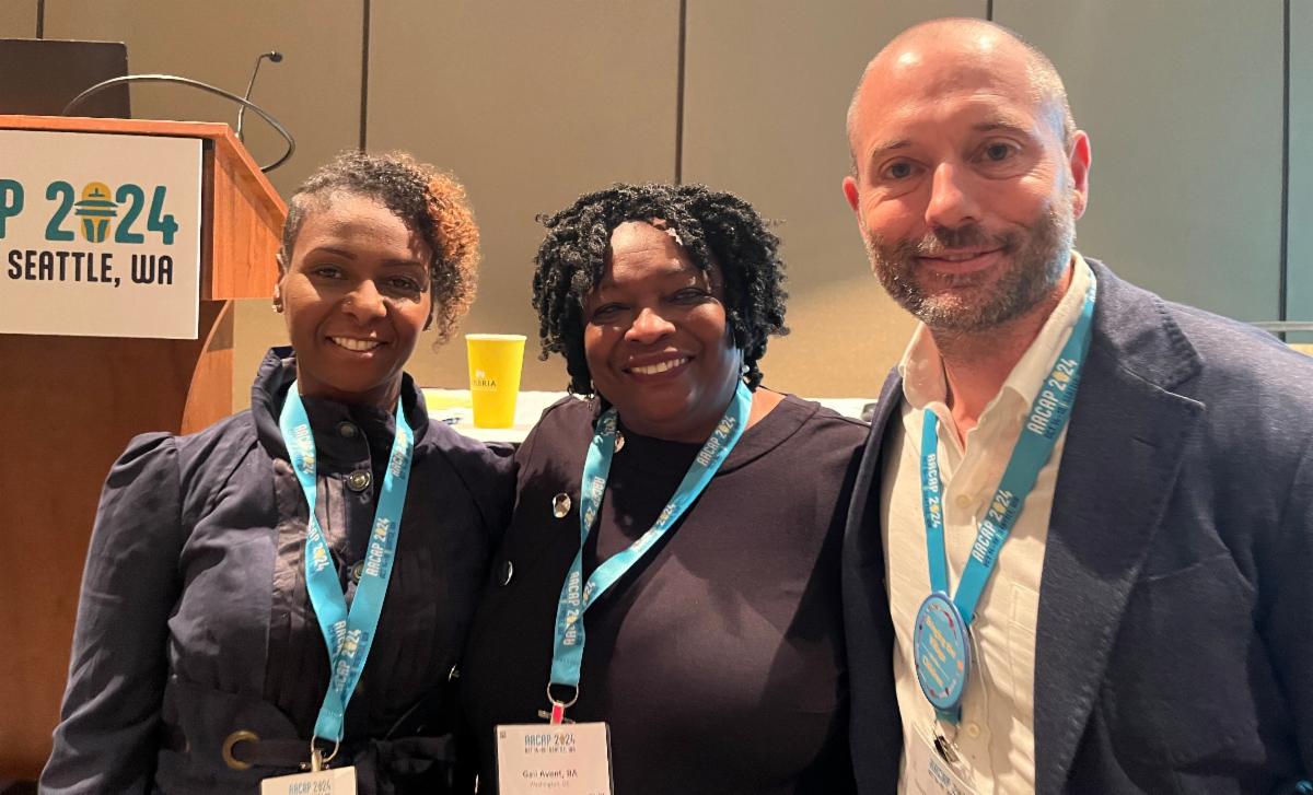 Three conference presenters wearing turquoise lanyards pose together at AACAP 2024 in Seattle, WA. In front of them is a wooden podium with the conference logo. All three presenters are smiling at the camera and dressed professionally, with two people in dark clothing and one person in a white shirt and dark jacket.