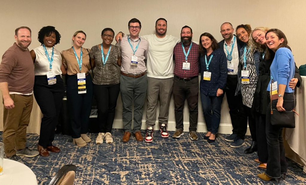 A group of eleven conference attendees stand together in a line against a beige wall, wearing conference lanyards with blue ribbons. The group is dressed in casual business attire, with some in sweaters and others in button-downs. They're standing on a blue patterned carpet and all are smiling warmly at the camera in a relaxed, friendly pose. AACAP 2024 in Seattle, WA.