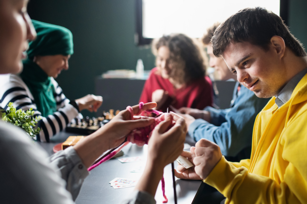 A group of young people gathered around a table in what appears to be a community or recreational space. In the foreground, someone in a bright yellow hoodie is looking at playing cards, while others are engaged in various activities. One person wears a green beanie hat and striped shirt, and there are craft materials or games visible on the dark table surface. The setting has a casual, social atmosphere with natural lighting coming from a window in the background.