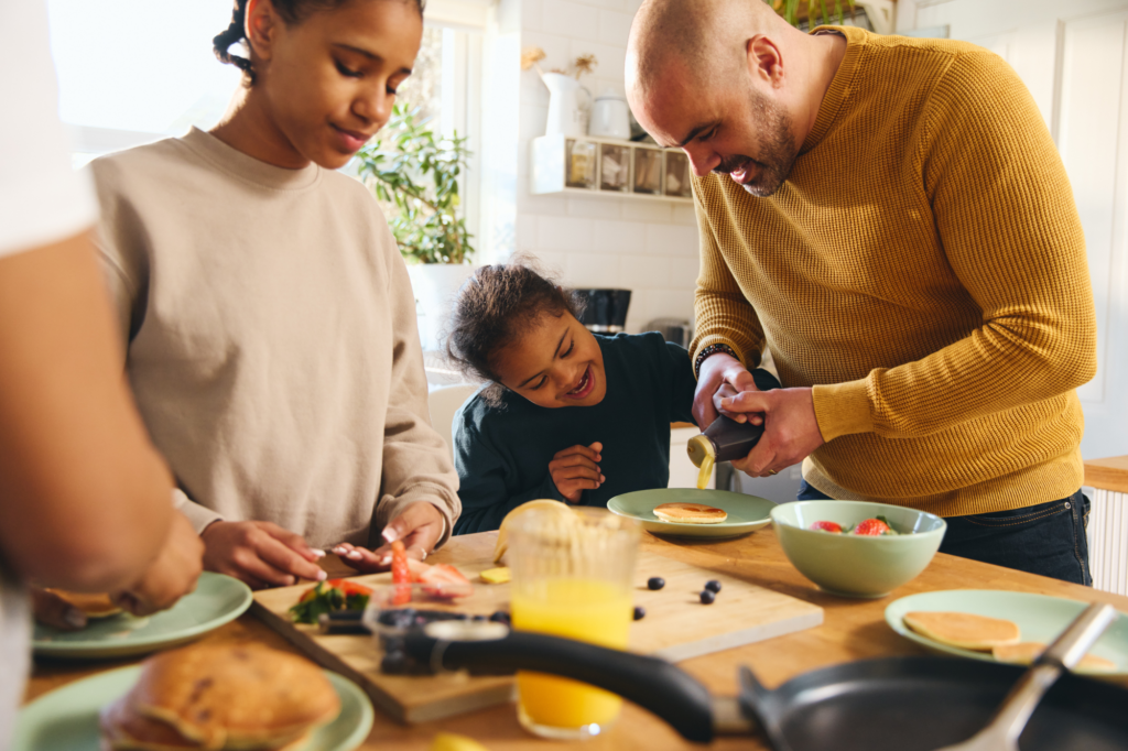 A warm family breakfast scene in a sunny kitchen. A father wearing a yellow sweater pours syrup on pancakes while his child laughs joyfully. A young person in a beige sweatshirt helps prepare food at the wooden table, which is set with green plates, berries, and orange juice. The image captures a moment of connection and happiness during a shared meal.