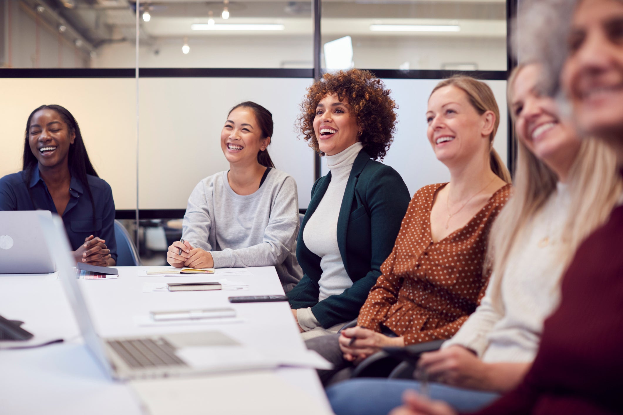 Line of women sitting around a large white table smiling and listening to a presentation that is off-camera.
