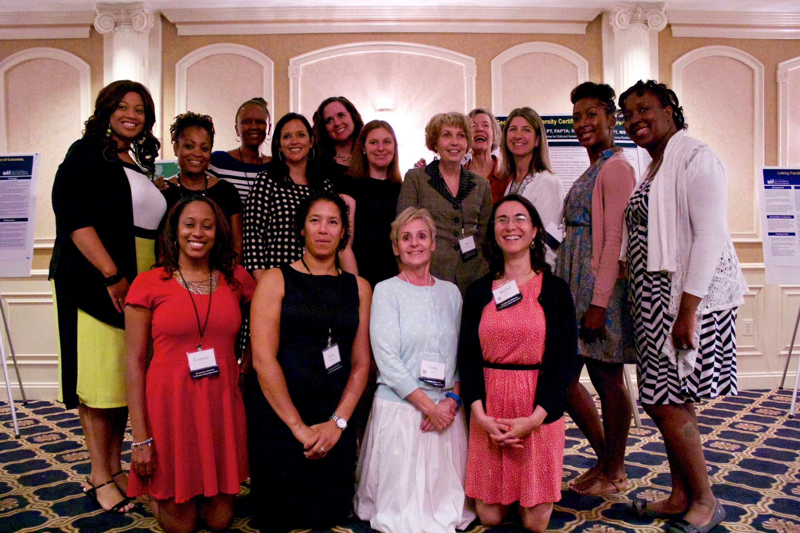 A group of fourteen women pose together at what appears to be a graduation or certificate ceremony, standing in front of a cream-colored wall with decorative moldings. They are arranged in two rows and dressed professionally in various outfits including dresses, blazers and business wear in colors like red, black, white and polka dots. Some wear name badges. Behind them are visible what appear to be academic or informational posters mounted on the wall.