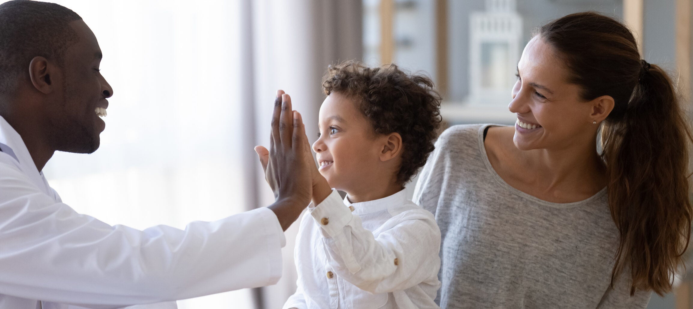 Happy african american young male doctor giving high five to smiling mixed race little kid boy, celebrating good result of prescribed medical treatment or welcoming new patient with mum at clinic.