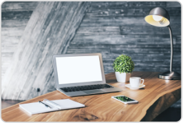 Modern home office workspace featuring a white laptop computer on a wooden desk with a steaming white coffee mug, small potted plant, notepad, and adjustable desk lamp against a textured blue-gray brick wall background.
