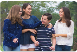 A family of four poses together outdoors with green foliage in the background. The group includes two adults and two children - a woman in a blue top with her arm around a young boy in a striped shirt, alongside another woman in a white top and a young girl. They are standing close together in a casual, friendly pose, suggesting a warm family moment captured in a natural outdoor setting.