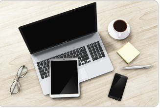 A laptop computer with a black screen sits open on a light wooden desk surface, accompanied by a white tablet, a black smartphone, a white coffee cup on a saucer, a small stack of yellow sticky notes, and a pair of eyeglasses. The items are arranged in a typical workspace setup, suggesting a modern office or home work environment.