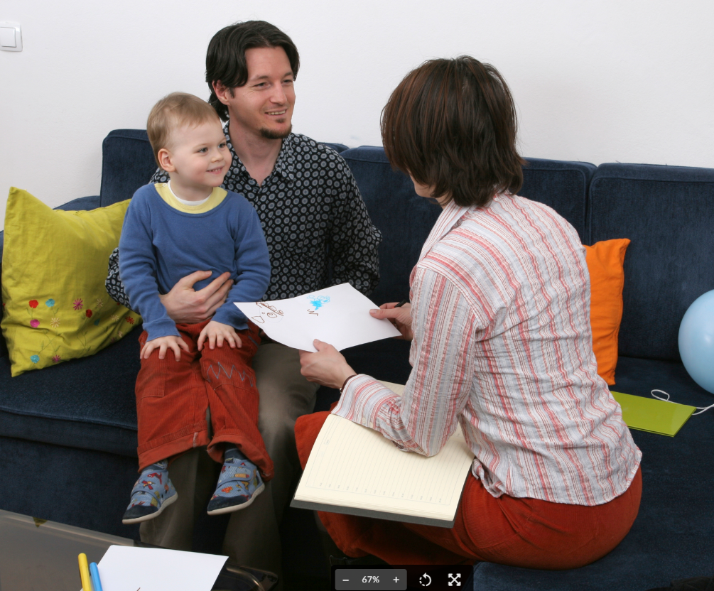 A father and young child sit on a blue couch while a woman in a striped shirt shows them a drawing on paper, appearing to be in a consultation or meeting setting.
