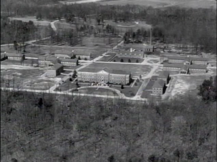 This black and white aerial photograph shows a large institutional complex with multiple rectangular buildings arranged around courtyards, surrounded by open fields and scattered trees in a rural setting.