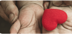 Close-up of elderly hands with wrinkled skin gently holding a small red heart-shaped object, likely fabric or felt, against a soft beige or tan colored background.