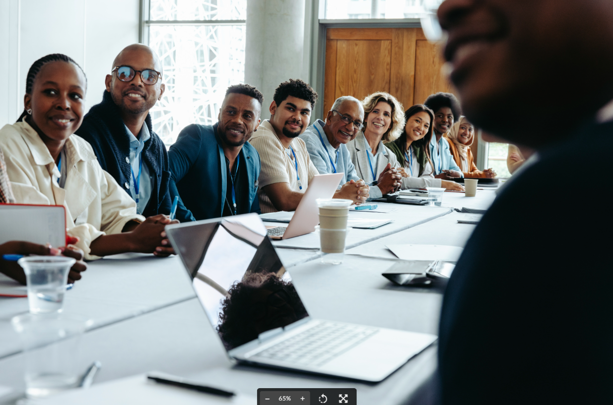 A diverse group of professionals with name badges sits smiling around a conference table during what appears to be a business meeting or training session, with laptops, notebooks, and coffee cups visible on the table.