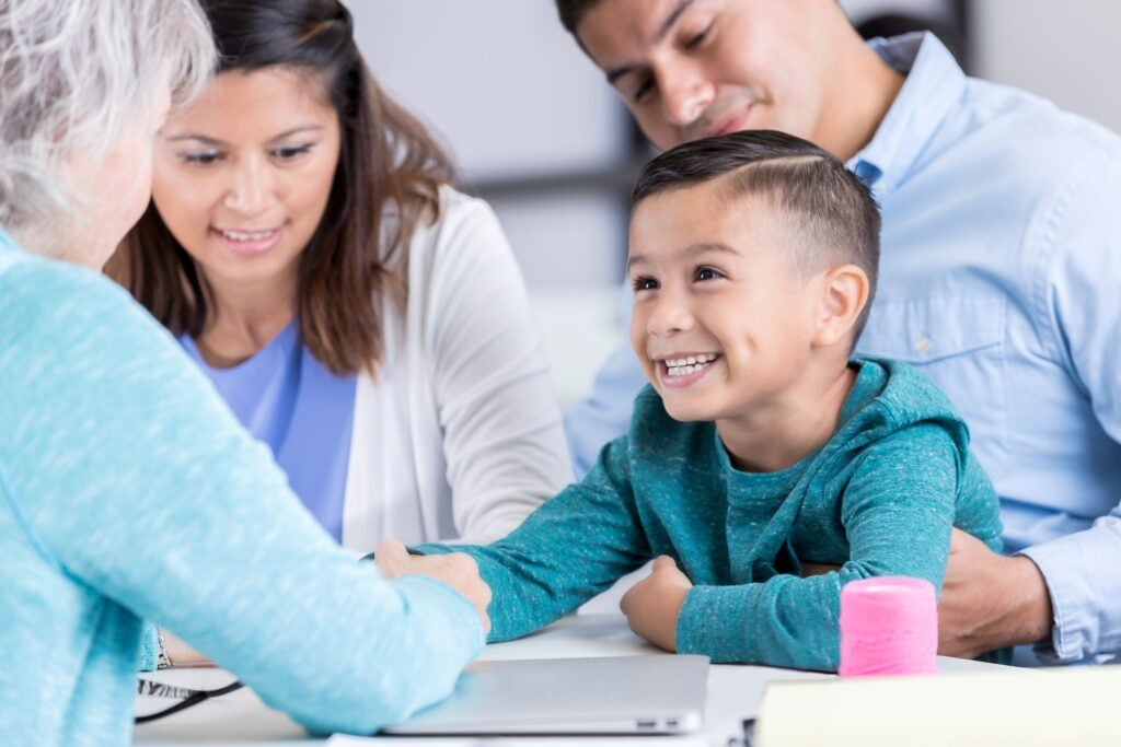 A smiling young boy in a teal sweatshirt sits at a table with three adults, who look on warmly.